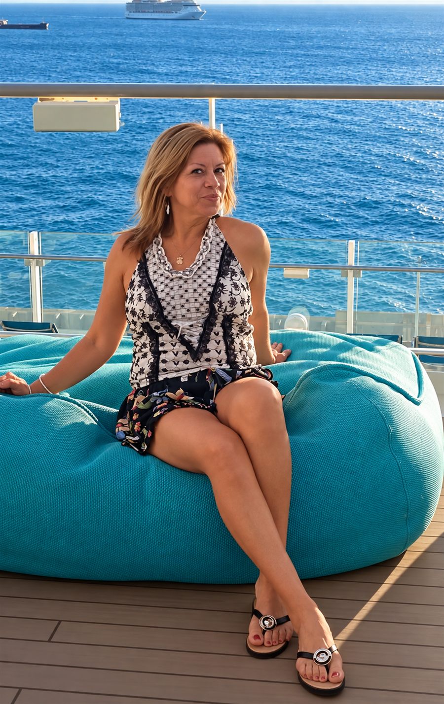 Marta sentada en un sillón turquesa junto al mar, con un crucero al fondo
